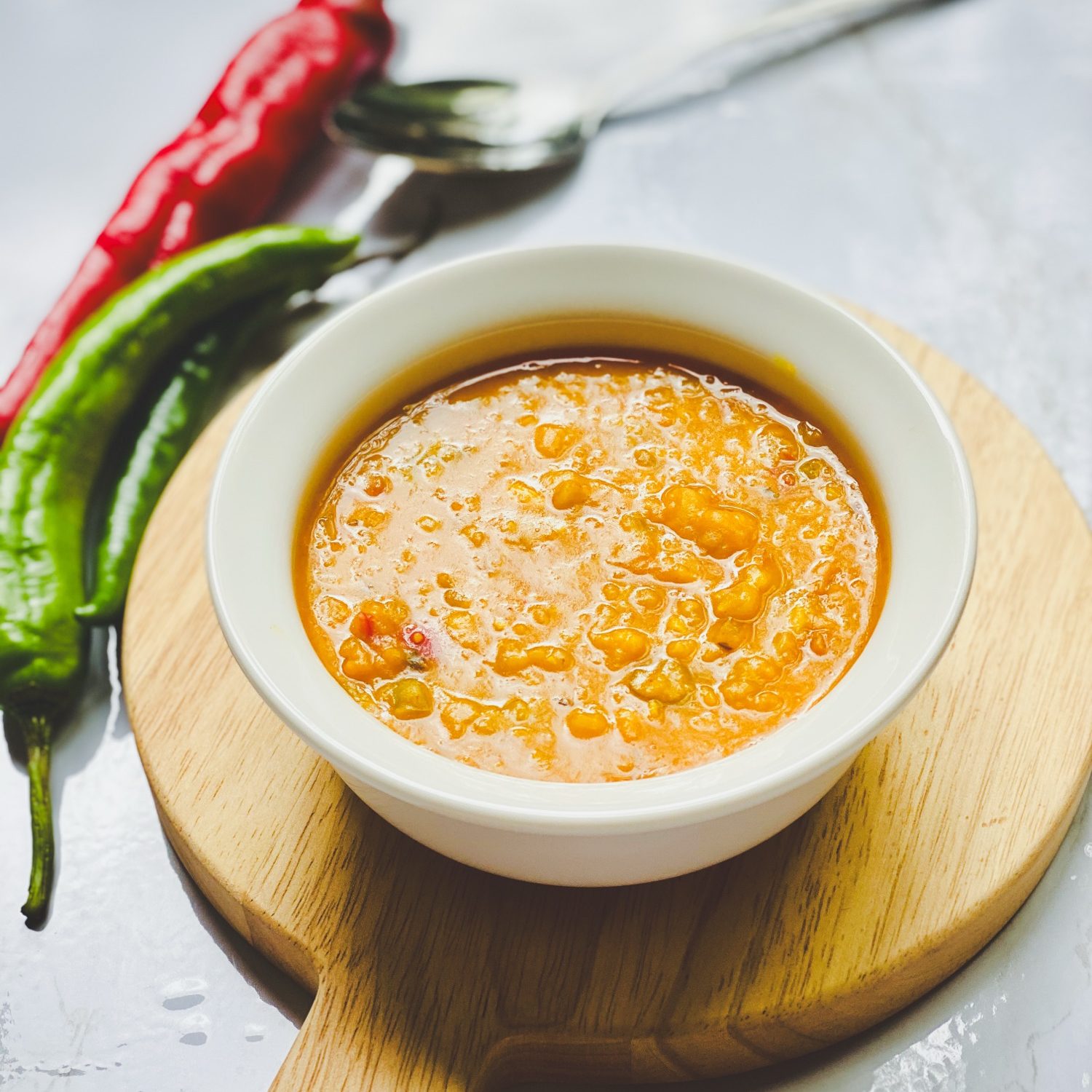 A bowl of daal in a white ceramic bowl placed on top of a round cutting board, with a red and green pepper positioned on the upper left corner of the image. A silver spoon is placed adjacent to the papers.